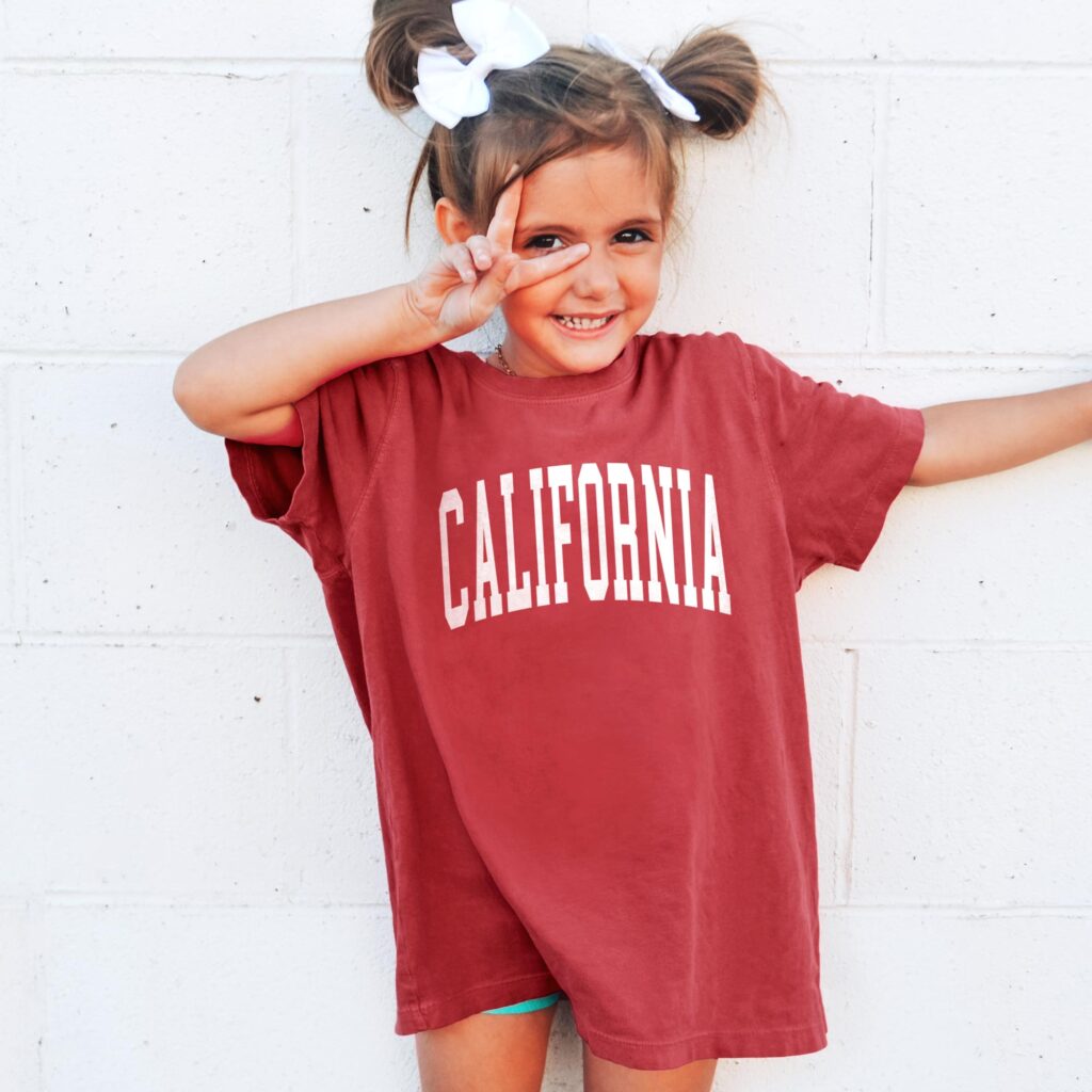 Young girl smiling and giving peace sign wearing red tee that says California.